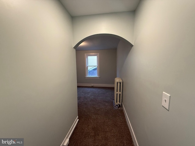 hall featuring arched walkways, radiator, baseboards, and dark colored carpet