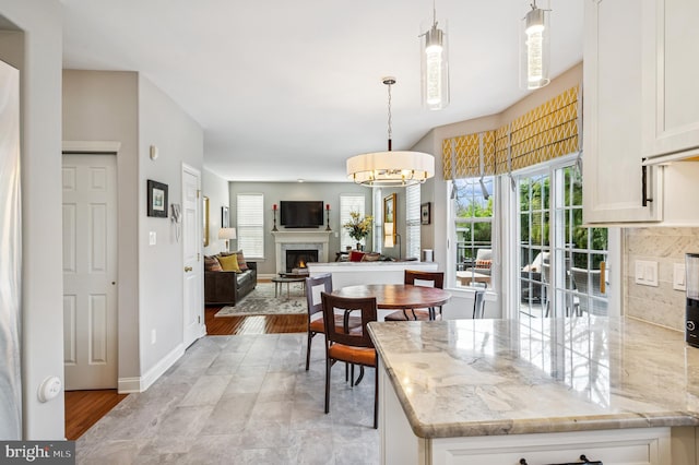 kitchen featuring pendant lighting, light stone counters, a warm lit fireplace, white cabinetry, and decorative backsplash