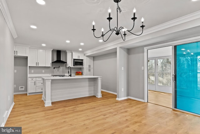 kitchen with ornamental molding, stainless steel microwave, wall chimney exhaust hood, light countertops, and a chandelier