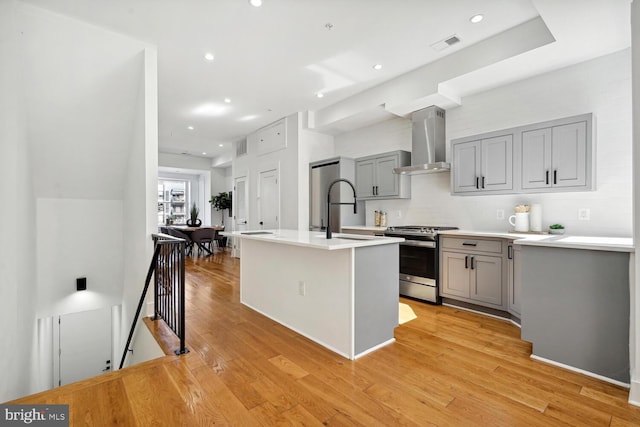 kitchen with light wood finished floors, gray cabinetry, wall chimney range hood, stainless steel range with gas stovetop, and a sink