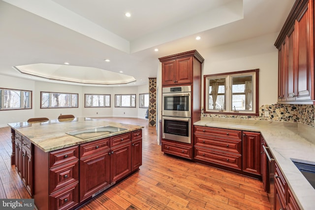 kitchen featuring a tray ceiling, light wood-style flooring, double oven, black electric cooktop, and reddish brown cabinets