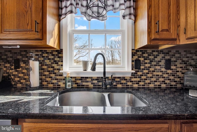 kitchen featuring decorative backsplash, dark stone countertops, brown cabinetry, and a sink