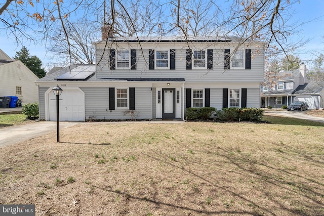 colonial inspired home with driveway, roof mounted solar panels, an attached garage, a front yard, and a chimney