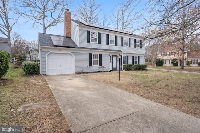 colonial-style house with solar panels, a chimney, concrete driveway, a front lawn, and a garage