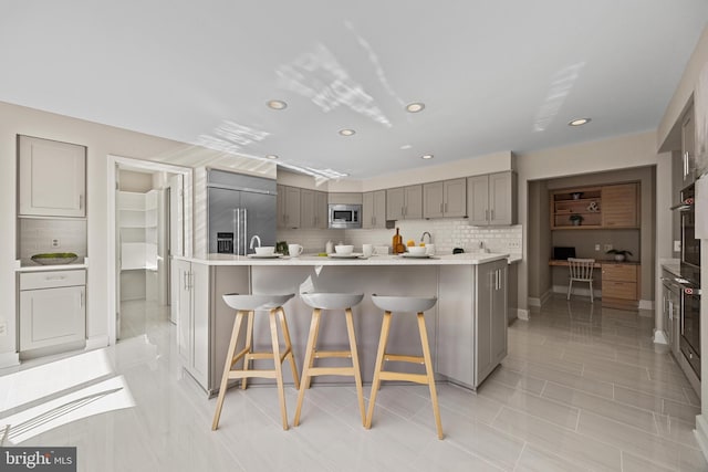 kitchen featuring built in appliances, a breakfast bar, light countertops, decorative backsplash, and gray cabinets