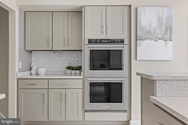 kitchen with a warming drawer, gray cabinets, stainless steel double oven, decorative backsplash, and light stone countertops