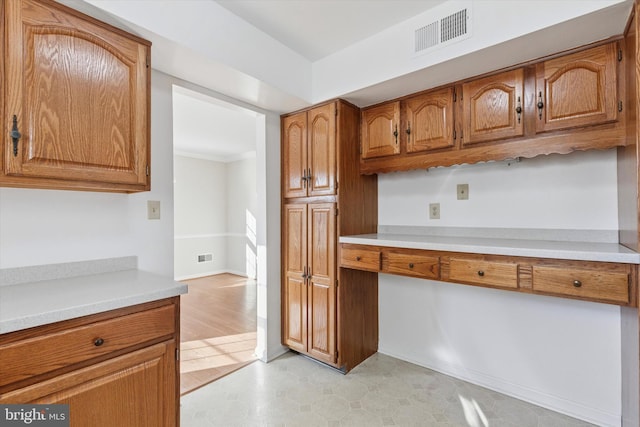 kitchen featuring visible vents, baseboards, light countertops, built in desk, and brown cabinetry