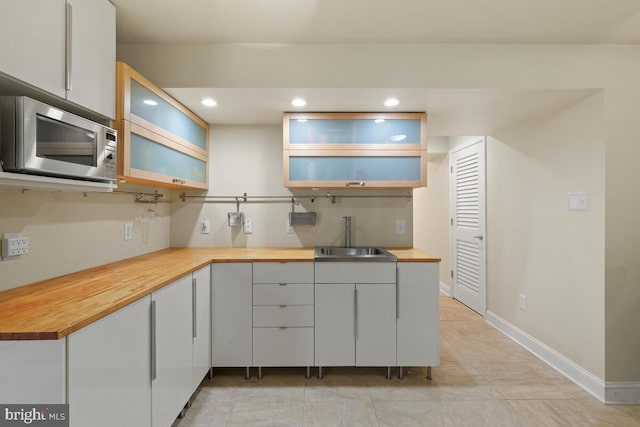 kitchen with baseboards, wooden counters, a sink, glass insert cabinets, and stainless steel microwave