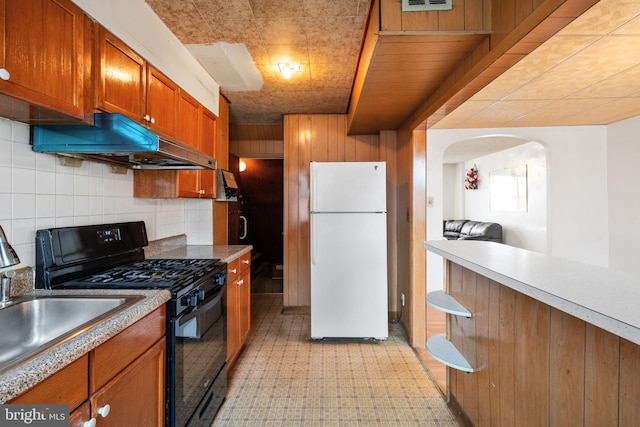 kitchen with brown cabinets, under cabinet range hood, tasteful backsplash, freestanding refrigerator, and black gas range oven