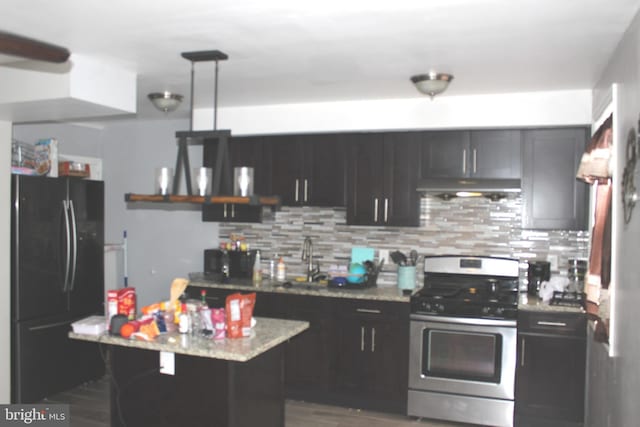 kitchen with stainless steel gas stove, under cabinet range hood, light stone counters, tasteful backsplash, and freestanding refrigerator