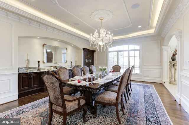 dining room with crown molding, an inviting chandelier, a decorative wall, a raised ceiling, and dark wood-style flooring