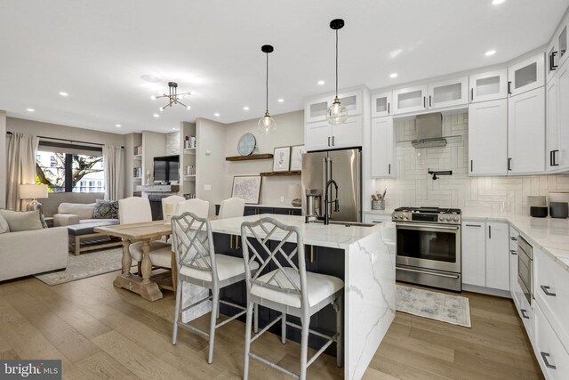 kitchen featuring light wood finished floors, tasteful backsplash, open floor plan, appliances with stainless steel finishes, and wall chimney exhaust hood