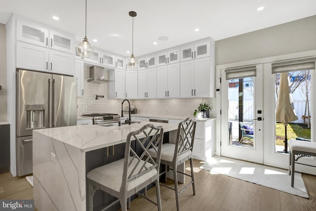 kitchen with light stone countertops, a center island with sink, stainless steel appliances, light wood-style floors, and wall chimney exhaust hood