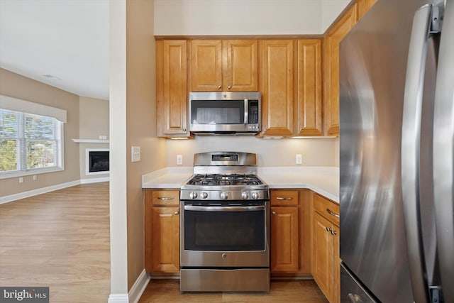 kitchen with baseboards, light wood-type flooring, light countertops, appliances with stainless steel finishes, and a glass covered fireplace