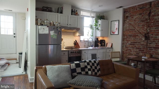 kitchen featuring tasteful backsplash, brick wall, under cabinet range hood, wood finished floors, and stainless steel appliances