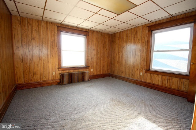 unfurnished room with a drop ceiling, wood walls, radiator, and carpet