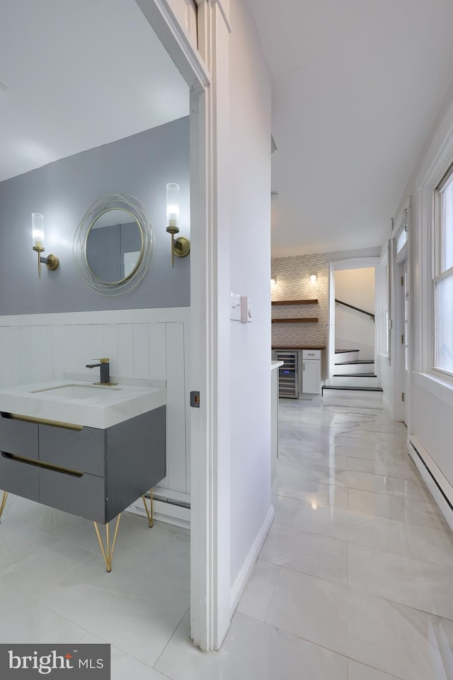 hallway featuring wine cooler, a wainscoted wall, stairs, baseboard heating, and a sink