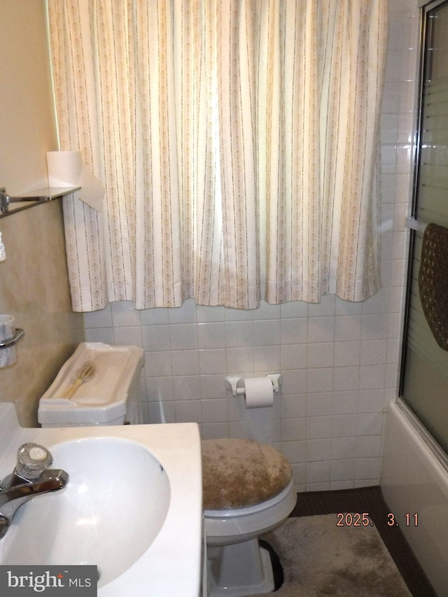 bathroom with tile walls, toilet, bath / shower combo with glass door, and a sink