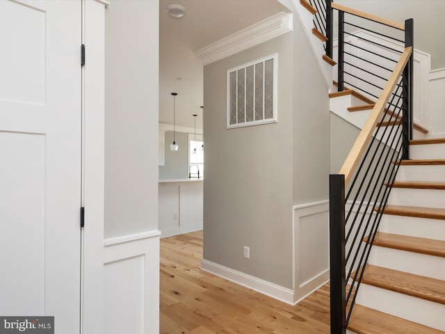 stairway with visible vents, wood finished floors, crown molding, and a decorative wall