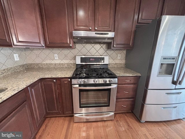 kitchen featuring under cabinet range hood, backsplash, stainless steel appliances, light wood finished floors, and light stone countertops