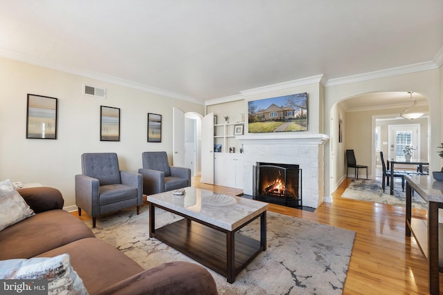 living area featuring visible vents, light wood-style flooring, a fireplace with flush hearth, arched walkways, and ornamental molding