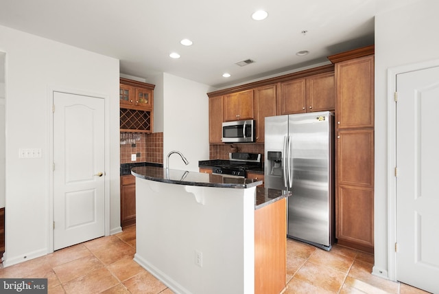 kitchen with brown cabinets, a center island with sink, stainless steel appliances, dark stone counters, and decorative backsplash