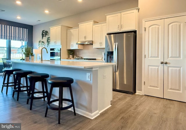 kitchen featuring a kitchen island with sink, stainless steel appliances, white cabinets, under cabinet range hood, and a kitchen bar