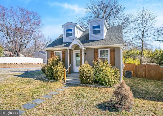 cape cod home with central AC unit, a shingled roof, a front lawn, and fence