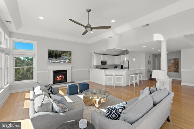 living room with visible vents, ornate columns, a tray ceiling, a lit fireplace, and light wood-type flooring