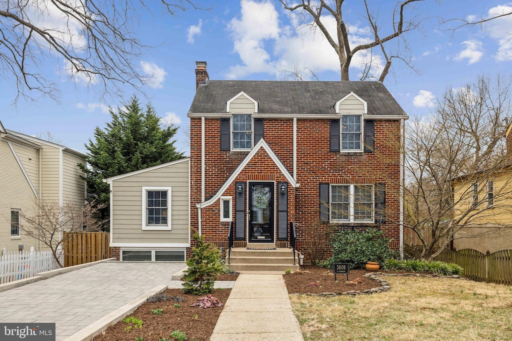 view of front of house featuring decorative driveway, brick siding, a chimney, and fence