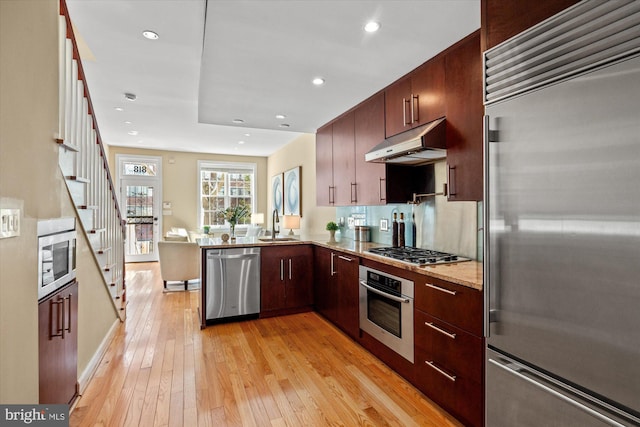 kitchen with under cabinet range hood, light wood-style flooring, appliances with stainless steel finishes, a peninsula, and a sink