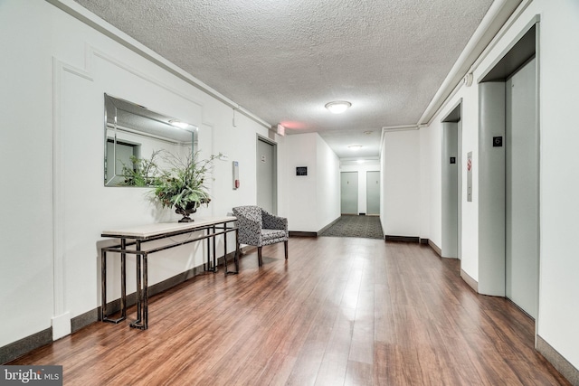 hallway with elevator, wood finished floors, baseboards, and a textured ceiling