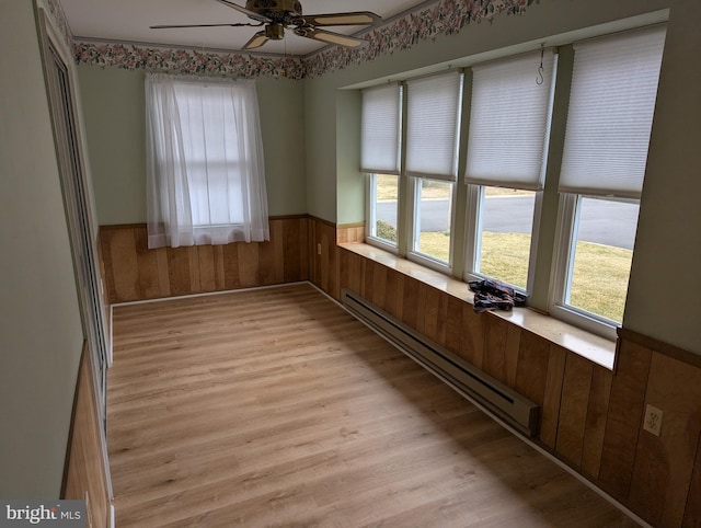 unfurnished dining area featuring wooden walls, a ceiling fan, light wood-style floors, wainscoting, and baseboard heating