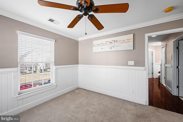 carpeted empty room featuring a wainscoted wall, crown molding, visible vents, and ceiling fan