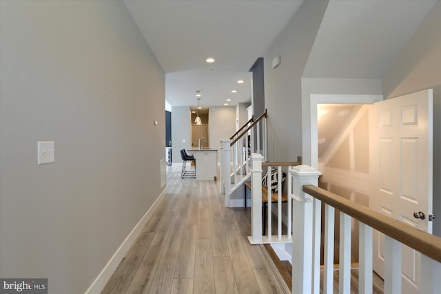 corridor featuring baseboards, light wood finished floors, recessed lighting, a sink, and stairs