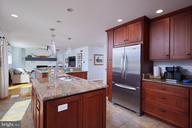 kitchen with light stone counters, a kitchen island with sink, a sink, stainless steel refrigerator, and open floor plan