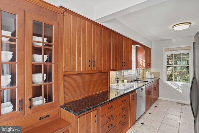 kitchen featuring tasteful backsplash, stainless steel dishwasher, light tile patterned flooring, brown cabinetry, and a sink