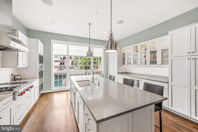 kitchen featuring an island with sink, a sink, stainless steel gas stovetop, white cabinetry, and wall chimney range hood