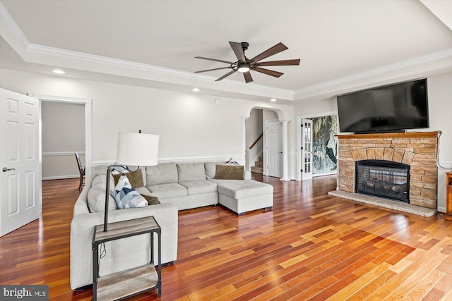 living room with ceiling fan, a tray ceiling, a fireplace, wood finished floors, and arched walkways