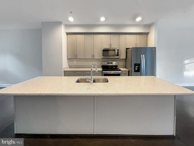 kitchen featuring light stone counters, a kitchen island with sink, a sink, gray cabinetry, and appliances with stainless steel finishes