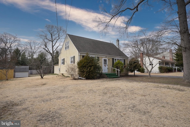 cape cod house with an outbuilding, fence, a storage shed, a shingled roof, and a chimney