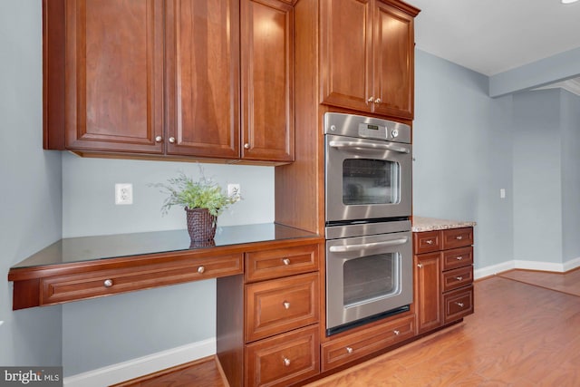 kitchen with stainless steel double oven, light wood-style flooring, brown cabinets, and baseboards