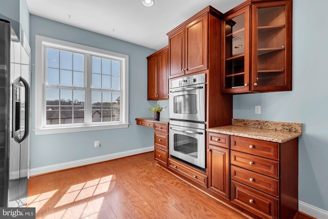 kitchen featuring light wood-type flooring, light stone counters, appliances with stainless steel finishes, glass insert cabinets, and baseboards