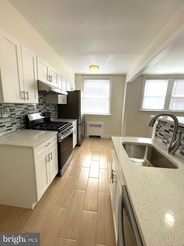 kitchen with radiator heating unit, a sink, stainless steel appliances, under cabinet range hood, and backsplash