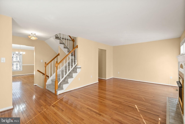 unfurnished living room featuring baseboards, stairs, hardwood / wood-style flooring, a brick fireplace, and a chandelier