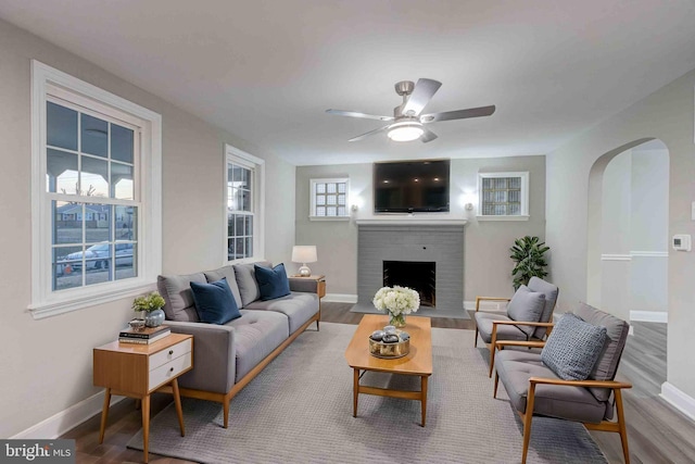 living room featuring ceiling fan, baseboards, a fireplace, wood finished floors, and arched walkways