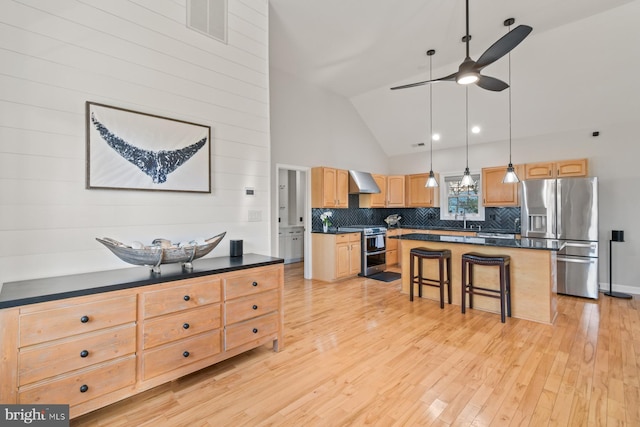 kitchen with dark countertops, appliances with stainless steel finishes, light brown cabinetry, and wall chimney range hood