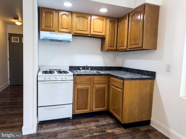 kitchen with a sink, white gas range oven, under cabinet range hood, dark countertops, and brown cabinets