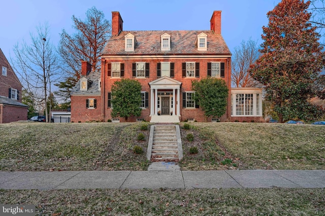 colonial-style house with a high end roof, a front yard, brick siding, and a chimney