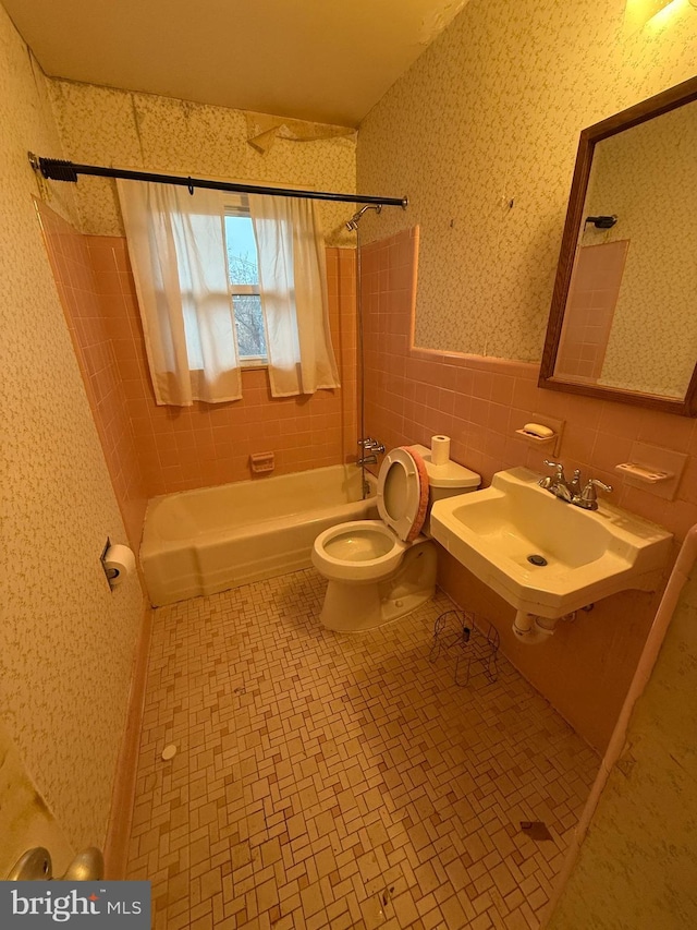 bathroom with wallpapered walls, toilet, bathing tub / shower combination, tile walls, and a sink